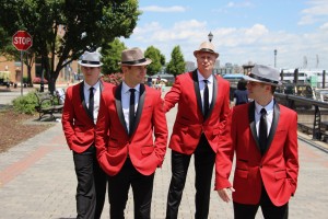 The Jersey Tenors singing group are shown walking down a street in red jackets and white hats.